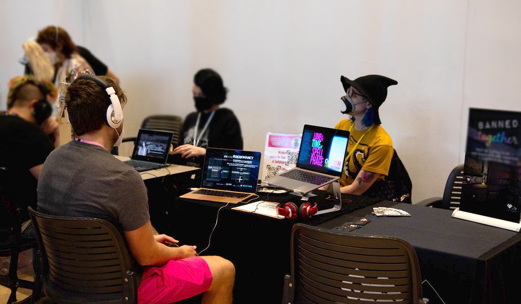 Official QGCon photo. Mackenzie sits and plays Fateweaver while Kylie in a witch hat sits behind the table.